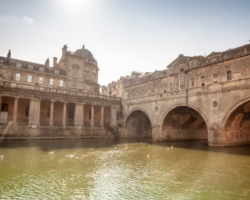 Historic buildings and arches reflect in calm waters under a bright sky, showcasing Bath's stunning architecture.