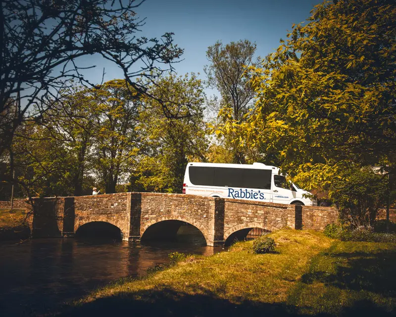 A Rabbie's Tours van crosses a picturesque stone bridge surrounded by lush greenery and a serene waterway.