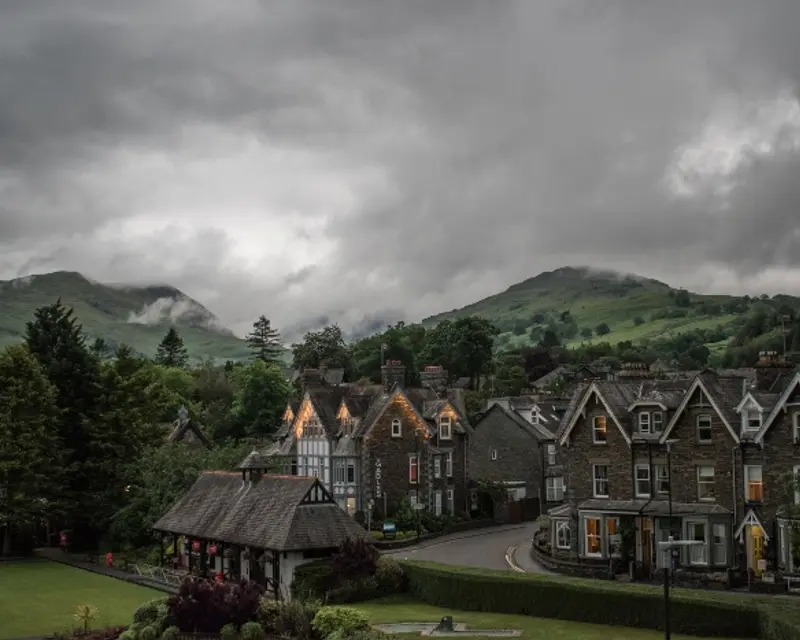 A quaint village with stone houses under a dramatic cloudy sky, surrounded by lush green hills.