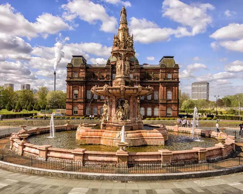 Victorian fountain in front of a grand building, surrounded by greenery and people, under a bright blue sky with fluffy clouds.