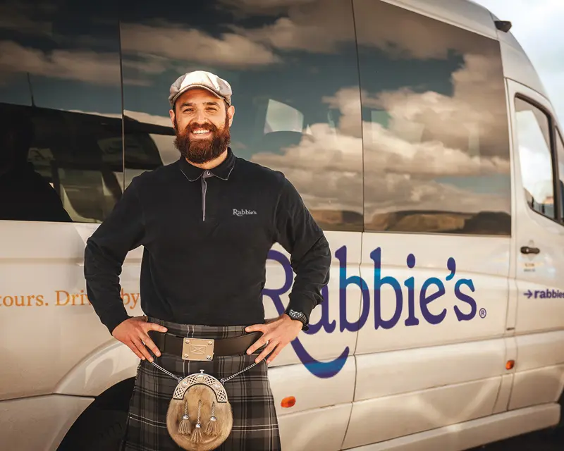 Smiling tour guide in a kilt stands in front of a Rabbie's Tours van, promoting small group tours in the UK.