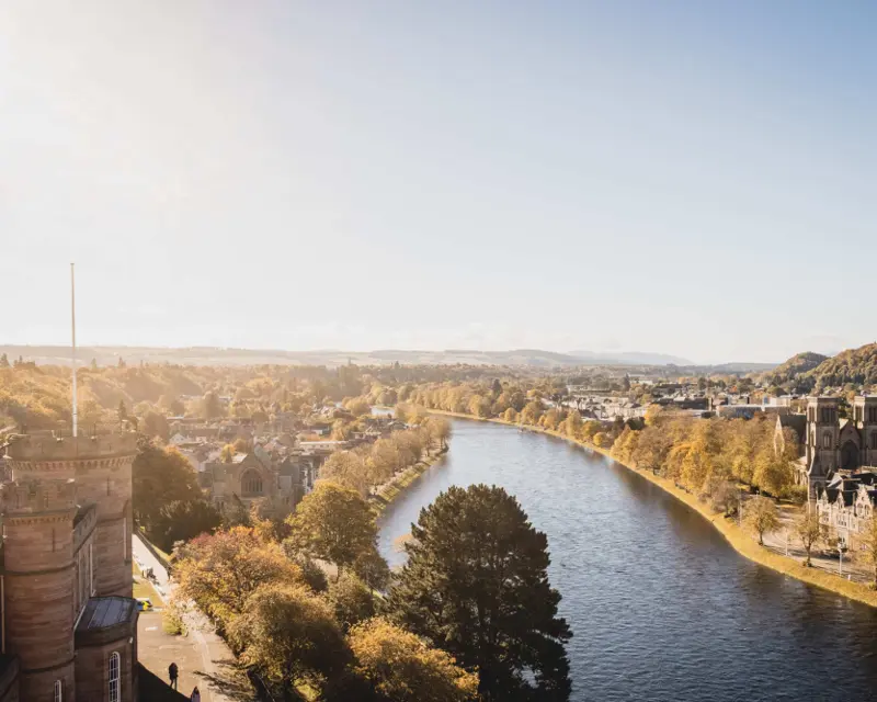 A scenic view of a river winding through autumn-colored trees and town buildings under a clear sky in the UK.