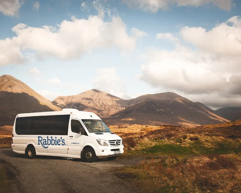 A Rabbie's Tours minibus parked on a scenic road with mountains and clouds in the background. Perfect for small group adventures.