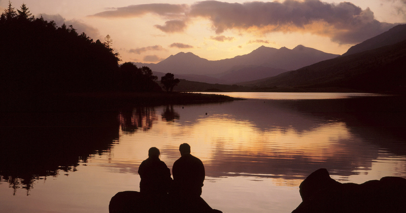 Two people sitting on a rock overlooking a lake at sunset