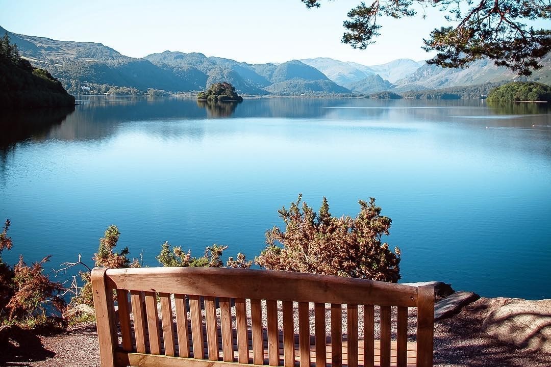 Lake District’s Derwentwater, brown bench in the foreground overlooking pale blue water surrounded by trees 