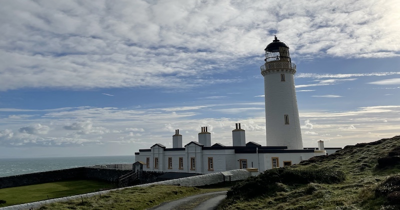 The tall, white Mull of Galloway lighthouse surrounded by green fields and overlooking an endless sea.