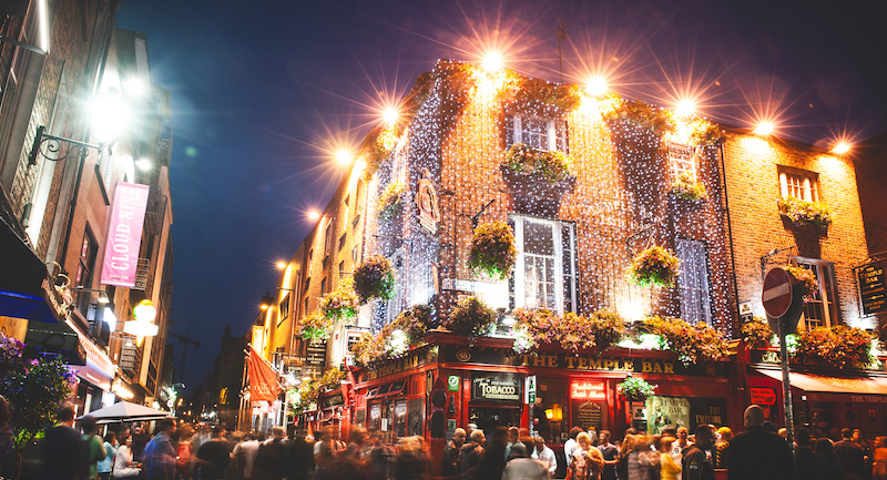 The famous Temple Bar in Dublin surrounded by crowds of people and lit up with fairy lights under a night sky.