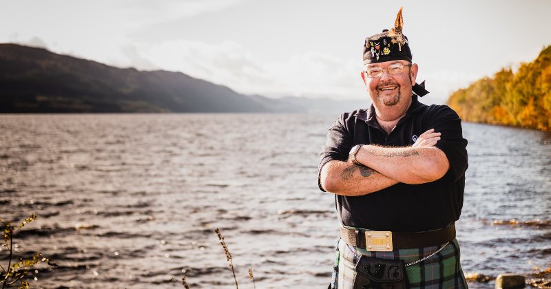 a Scottish clan member wearing a kilt and smiling in front of a loch