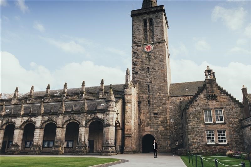 a stone building located in St Andrews featuring a red clock on the buildings tower and an arched walkway 