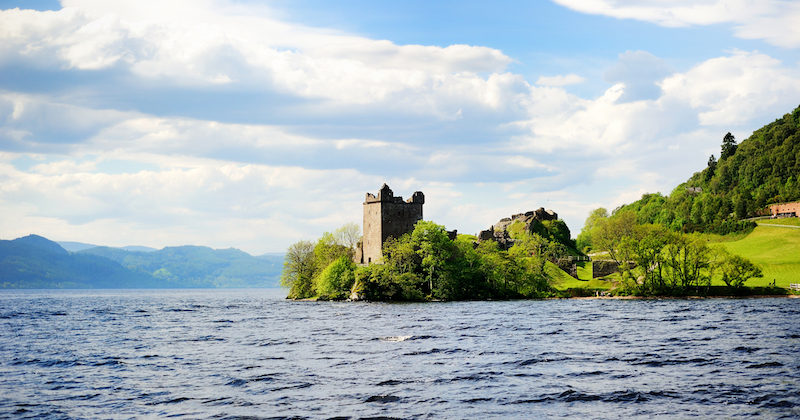 Urquhart Castle sits on the green banks of Loch Ness overlooking the clear, blue water in summer