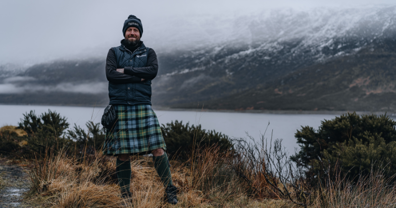 a man standing in a kilt with a loch behind him